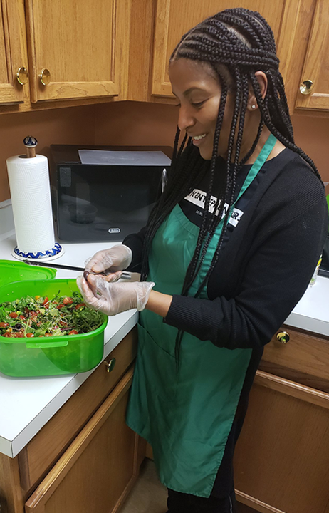 A smiling Black woman is assembling a salad in a small kitchen.